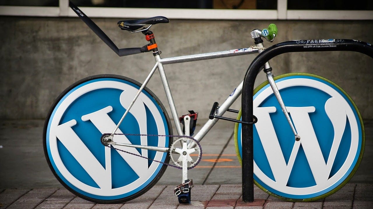 Ein Fahrrad ist an einem Ständer geparkt, seine Räder wurden durch große WordPress-Logos anstelle von Reifen ersetzt - die perfekte Inspiration, wenn Sie Die besten WordPress-Themes für 2024 finden wollen. Der silberne Rahmen des Fahrrads kontrastiert mit dem schwarzen Sitz und Lenker.