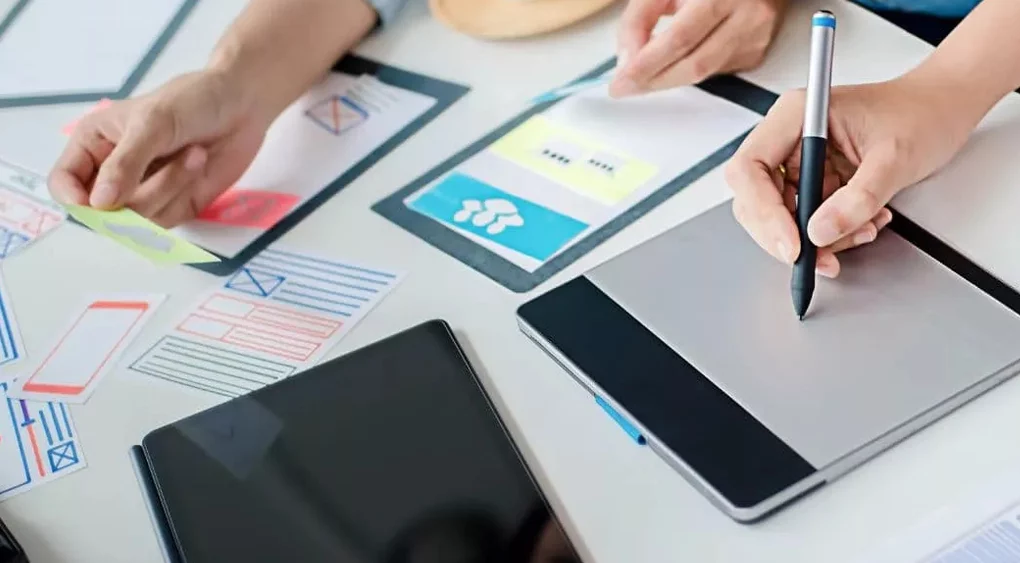 Zwei Personen arbeiten an einem Schreibtisch mit einem Tablet, einem digitalen Stift, Papierprototypen, Haftnotizen und einem Tablet und erarbeiten gemeinsam UI/UX-Designkonzepte für ein digitales Projekt, während sie die angesagtesten Webdesign-Trends des Jahres 2024 erkunden.
