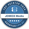 Eine blau-weiße kreisförmige Plakette mit dem Schriftzug Top Agentur 2024, JOMOX Media, fünf Sternen und agenturTIPP.de. Oben in der Mitte erscheint ein einfaches Gebäudelogo. Eine blau-weiße kreisförmige Plakette mit dem Schriftzug Top Agentur 2024, JOMOX Media, fünf Sternen und agenturTIPP.de. Oben in der Mitte erscheint ein einfaches Gebäudelogo.