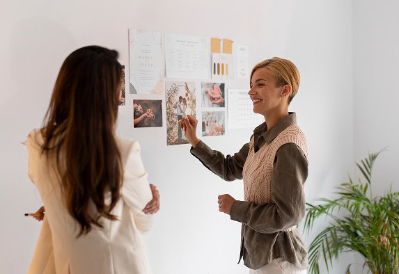 Zwei Frauen stehen an einer Wand, an die Fotos und Papiere gepinnt sind. Eine Frau lächelt und zeigt auf die Wand, während die andere ihr zugewandt ist und zuhört. In der Ecke des Raums steht eine Grünpflanze.