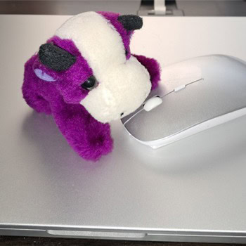 Ein kleines lila-weißes Stofftier, das einer Kuh ähnelt, ruht auf einer silbernen Computermaus, die auf einem silbernen Laptop steht.
