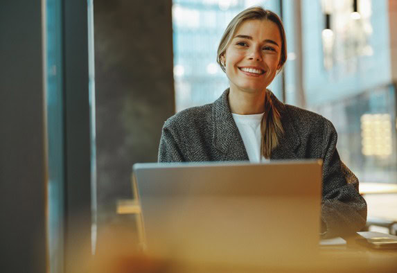 Eine junge Frau mit blonden Haaren, grauem Blazer und weißem Hemd, sitzt lächelnd mit ihrem Laptop an einem Tisch in einem modernen, sonnendurchfluteten Büro - perfekt für die Erledigung von Aufgaben, die den ganzen Tag über grundlegende Leistungen erfordern.