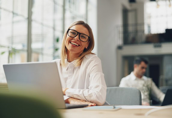 Eine Frau, die eine Brille und ein weißes Hemd trägt, lächelt, während sie an einem Schreibtisch mit einem Laptop sitzt und grundlegende Aufgaben in einem hellen, modernen Büro ausführt. Eine weitere Person ist unscharf im Hintergrund zu erkennen.