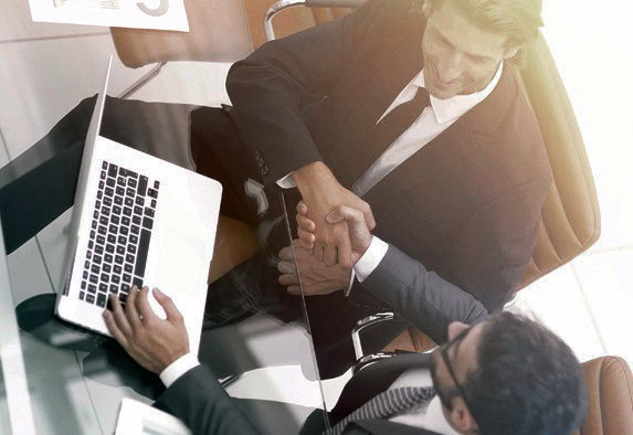 Zwei Geschäftsleute in Anzügen sitzen an einem gläsernen Schreibtisch mit einem Laptop, schütteln sich die Hände und lächeln, von oben betrachtet, was auf eine erfolgreiche Geschäftsvereinbarung oder -besprechung schließen lässt.