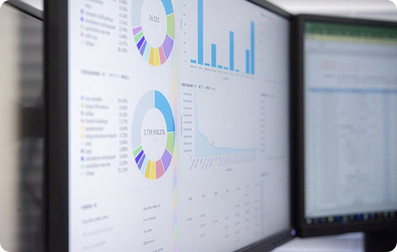 Zwei Computermonitore zeigen Datenvisualisierungen an, darunter bunte Kreisdiagramme, Balkendiagramme und Tabellen. Die Bildschirme zeigen detaillierte Statistiken und analytische Informationen in einer hellen Büroumgebung.