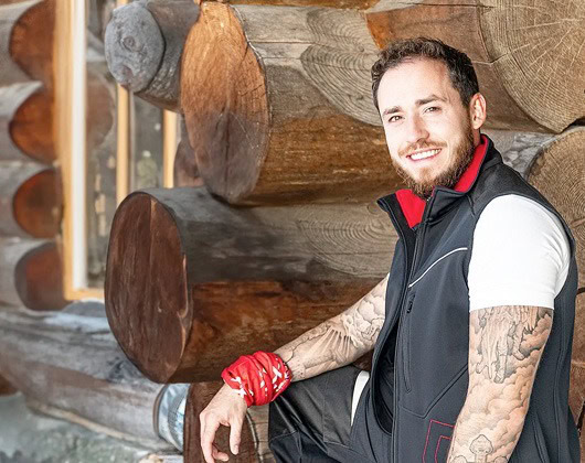 Ein lächelnder Mann mit Tätowierungen und einem roten Halstuch am Handgelenk sitzt vor einer Blockhütte und trägt eine schwarze Weste über einem weißen Hemd - ein lässiger Stil mit RG Arbeitsschutz-Flair.