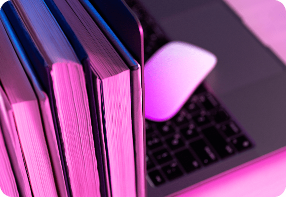 Drei dicke Bücher stehen aufrecht neben einem aufgeklappten Laptop und einer kabellosen Maus, alles beleuchtet von rosa und lila Licht.