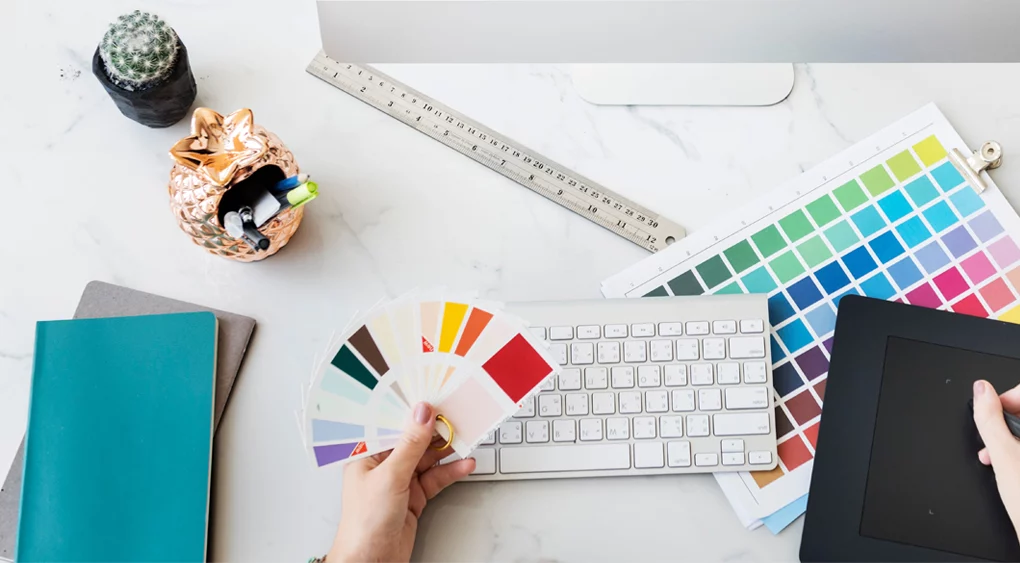 Eine Person hält Farbmuster über einer Tastatur auf einem Schreibtisch, neben einem Grafiktablett, Notizbüchern und einem Stifthalter - perfekt für Logo-Design oder Visitenkarten-Branding-Inspiration - auf einer Marmorfläche mit einem Lineal und einem kleinen Kaktus.