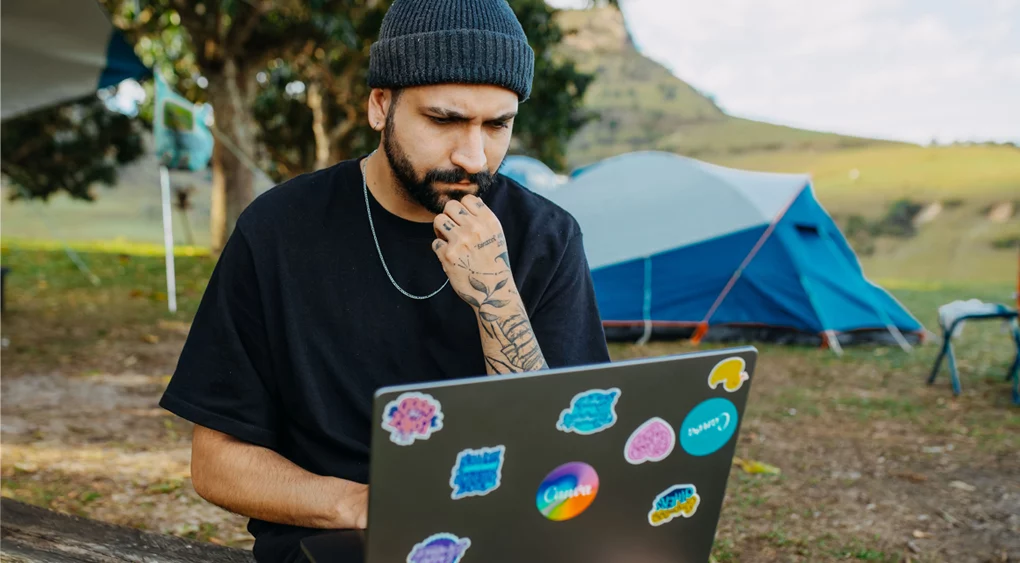 Eine Person mit Mütze und schwarzem T-Shirt sitzt im Freien und blickt nachdenklich auf einen Laptop, der mit bunten Aufklebern übersät ist – vielleicht sucht sie nach Ideen für ein Logo-Design. Zelte und Bäume im Hintergrund lassen auf eine kreative Campingatmosphäre schließen.
