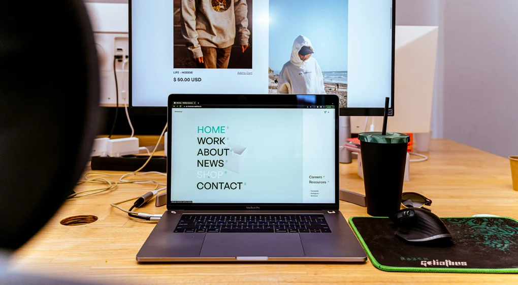 Auf einem Schreibtisch steht ein Laptop, auf dem eine Website angezeigt wird. Daneben liegen eine Maus, ein Mauspad, eine Tasse und im Hintergrund ist ein Monitor zu sehen, auf dem eine Bekleidungs-Website zu sehen ist. Auf der linken Seite des Schreibtisches sind Kabel und Steckdosen zu sehen, was auf subtile Weise den digitalen Arbeitsbereich hervorhebt, in dem für Online-Unternehmen oft Überlegungen wie „Landingpage vs. Website“ eine Rolle spielen.