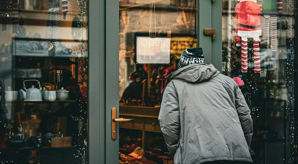 Eine Person in einem grauen Mantel und einer Wintermütze beugt sich vor, um durch das Schaufenster eines mit Lichterketten und Weihnachtsschmuck geschmückten Ladens zu schauen. Ein Schild an der Tür mit der Aufschrift „Geöffnet“ lädt Passanten ein und verdeutlicht, wie lokale SEO für Unternehmen lokalen Geschäften helfen kann, in der Weihnachtszeit mehr Kunden anzulocken.