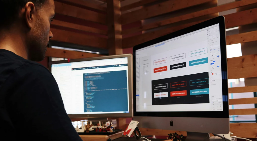 Eine Person arbeitet an einem Schreibtisch mit zwei Bildschirmen; auf dem einen wird Code angezeigt, auf dem anderen ein Designprojekt mit farbigen Textfeldern, die sich auf die SEO-Optimierung im Rahmen einer Website-Migration beziehen. Die Kulisse scheint ein modernes Büro mit Holzwänden zu sein.