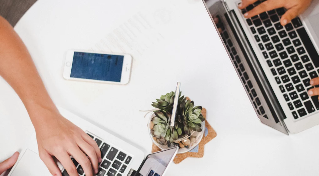 Zwei Personen, die an einem weißen Tisch an ihren Laptops arbeiten und über SEO für KMU diskutieren; in der Mitte stehen ein Smartphone und eine kleine Sukkulente im Topf.