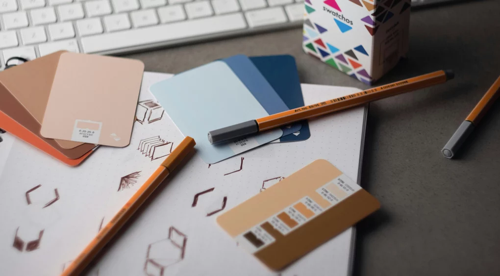 Ein Arbeitsplatz mit Farbmusterkarten, zwei orangefarbenen Stiften, einer Tastatur und einem Notizbuch mit geometrischen Skizzen – perfekt für alle, die ein Corporate Design erstellen möchten. Die Farbmusterkarten sind in Beige- und Blautönen gehalten, wobei einige davon bewusst auf dem Notizbuch platziert wurden, um Sie zu Ihrem nächsten kreativen Konzept zu inspirieren.