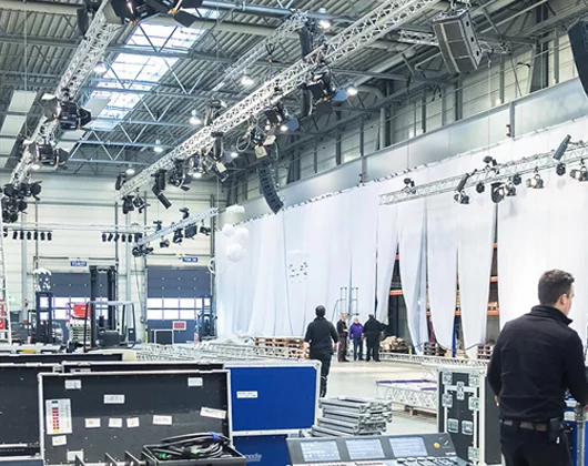 Großer Veranstaltungsraum mit Beleuchtungsanlagen, Vorhängen und Gerätekisten. Mehrere Personen richten den Raum ein und bereiten ihn für eine Multimedia-Veranstaltung oder -Show vor. Hohe Decken und industrielle Elemente sind zu sehen, was auf eine dynamische Umgebung hindeutet, die sich ideal für die Installation fortschrittlicher Mediensysteme eignet.